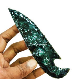 Puede incluir: Una punta de flecha de obsidiana verde oscuro con un patrón ondulado único. La punta de flecha se sostiene en una mano, mostrando su tamaño y detalles.
