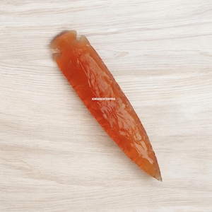 May include: A large, orange-red, arrowhead-shaped piece of obsidian. The obsidian is smooth and polished, with a slight sheen. The obsidian is lying on a light wood surface.