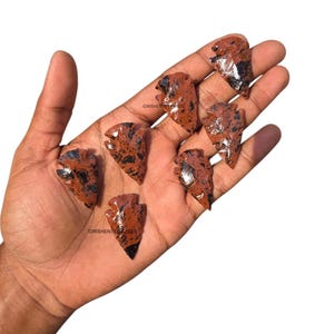 May include: Seven red-brown obsidian arrowheads with black speckles are displayed. The arrowheads are of varying sizes and shapes, with sharp points and detailed edges. The image is set against a white background.