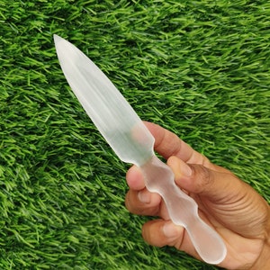 Puede incluir: Un cristal de selenita transparente con forma de cuchillo. El cristal es liso y pulido, con una punta afilada y un mango redondeado.