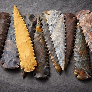 May include: Seven stone arrowheads with jagged edges are displayed. The arrowheads are in various colours, including brown, grey, and yellow. The image is set against a dark grey background, with the text "IDRISH ENTERPRISES" visible.