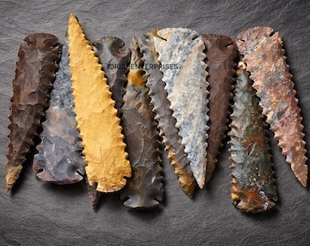 May include: A collection of seven stone arrowheads with serrated edges. The arrowheads vary in color, including shades of brown, gray, and yellow. The image is set against a dark gray background, with the text "IDRISH ENTERPRISES" visible.