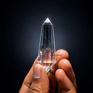 以下が含まれることがあります： 透明でファセット加工されたクリスタルオベリスク。尖った先端が特徴です。暗い背景に、手でクリスタルが持たれています。オベリスクは透明で、光を通し、研磨された外観をしています。クリスタルは装飾品です。