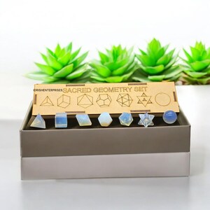 May include: A set of seven clear, iridescent, geometric shapes in a wooden box. The box has the words "Sacred Geometry Set" printed on it. The shapes include a pyramid, cube, tetrahedron, octahedron, dodecahedron, icosahedron, and a Star of David.