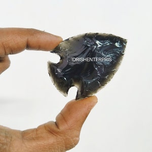 Puede incluir: Una punta de flecha gris oscuro con una base con muescas, sostenida entre los dedos. La punta de flecha tiene una punta afilada y una superficie texturizada. El texto "IDRISHENTERPRIS" es visible en la punta de flecha.