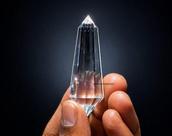 May include: A clear, faceted, double-terminated quartz crystal point with a pointed top and bottom. The crystal is transparent with visible internal inclusions and features the small text IDRISHENTERPRISES printed on its surface.