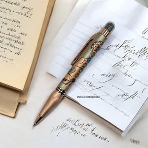 May include: A decorative pen with a rose gold finish and intricate floral and geometric designs. The pen features a clear, textured top and is lying on a white surface with handwritten notes and a book.