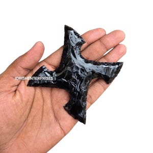 Puede incluir: Una piedra de obsidiana negra con forma de estrella o cruz de cuatro puntas, con bordes afilados y dentados y una superficie brillante y reflectante. El texto IDRISHENTERPRISES está impreso en blanco en el lado izquierdo del objeto oscuro.