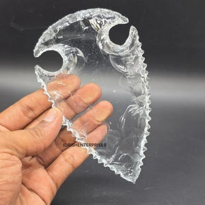 May include: A clear, translucent arrowhead-shaped object with a serrated edge and three curved openings. The text "IDRISHENTERPRISES" is visible. The object is held in a hand against a dark background.