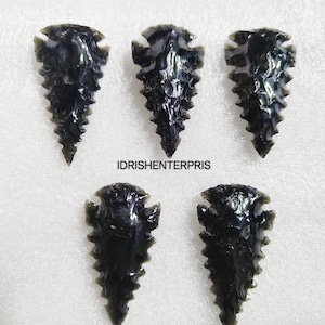Puede incluir: Cinco puntas de flecha de obsidiana negra con un borde dentado. Las puntas de flecha están dispuestas en fila sobre una superficie blanca.