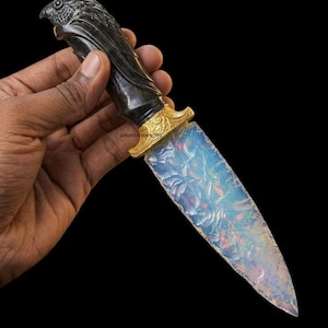 May include: A decorative knife featuring a translucent, iridescent opal-like blade with a textured surface. The handle is carved from dark material into the shape of a bird head, and a gold-colored metal bolster with intricate scrollwork connects it.
