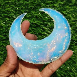 Pode incluir: Uma peça de opala em forma de lua crescente com uma cor azul e iridescente. A opala é polida e tem uma superfície lisa.