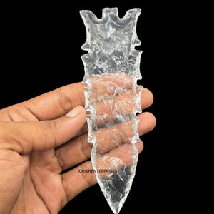 May include: A clear, transparent arrowhead with a textured surface and a pointed tip. The arrowhead is held against a black background. The text "IDRISHENTERPRISES" is visible near the bottom of the image.