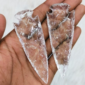 May include: Two clear quartz arrowheads, also known as arrow points, are held in a hand. The arrowheads are translucent and have a smooth, polished surface.