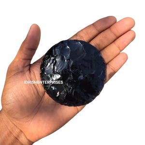 Puede incluir: Una piedra de obsidiana negra pulida y redonda. La piedra se sostiene en una mano, mostrando su superficie lisa y reflectante. El texto "IDRISHENTERPRISES" es visible. Parece ser un espécimen geológico natural.