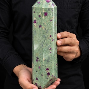 Op de afbeelding: Een gepolijste zeshoekige kristaltoren met een lichtgroene basissteen met ingebedde diepmagenta en donkerrode minerale insluitingen over het hele oppervlak, eindigend in een scherpe, veelzijdige punt aan de bovenzijde.