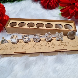 May include: A wooden box with eight compartments for storing geometric crystal shapes. The box has a label that reads "Sacred Geometry". The crystals are clear and include a sphere, a pyramid, a cube, and a star.