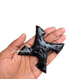 May include: A black obsidian arrowhead with a four-point design. The arrowhead is held in a hand against a white background. The text "IDRISHENTERPRISES" is visible on the image.