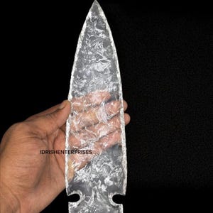 May include: A clear, glass arrowhead with a textured surface and a pointed tip. The arrowhead has a rectangular base with two curved cutouts. The text "IDRISHENTERPRISES" is visible on the arrowhead.