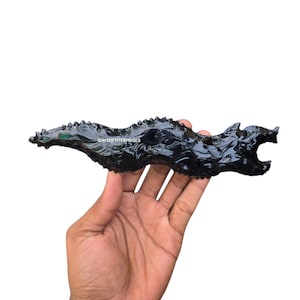 Puede incluir: Una talla de obsidiana negra con forma de dragón, sostenida en una mano. El dragón tiene una superficie texturizada y detalles. La talla mide aproximadamente 25 cm de largo.