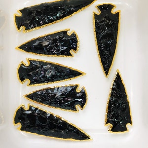 May include: Six black obsidian arrowheads with gold trim. The arrowheads are arranged in a row on a white surface.