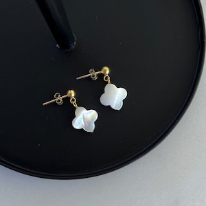 May include: A pair of gold earrings with white shell flower-shaped charms. The earrings are displayed on a black surface.