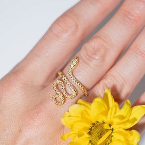 May include: A gold snake ring with a textured design. The ring is worn on a finger with a yellow flower.