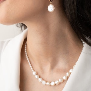 May include: A white pearl necklace with a simple design. The necklace is made of small, round pearls that are strung together on a thin chain. The necklace is worn by a person with long, dark hair.