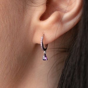 May include: A silver hoop earring with small, clear stones along the hoop and a teardrop-shaped, purple gemstone pendant. The earring is worn in an ear, with the skin visible in the background.