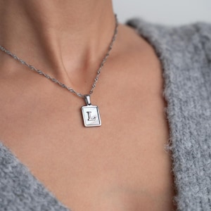 May include: A silver chain necklace with a silver pendant featuring the letter "L" in a square frame.