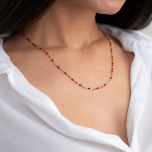 Peut inclure: Un collier en chaîne dorée avec de petites perles rouges. Le collier est porté par une personne aux cheveux foncés et à la chemise blanche.