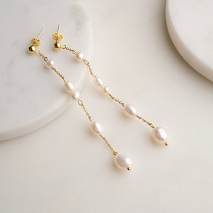 May include: A pair of long, gold chain earrings with white pearl accents. The earrings are on a white surface.
