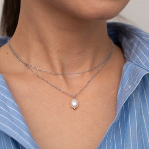 May include: A silver chain necklace with a single pearl pendant. The necklace is layered with two chains.