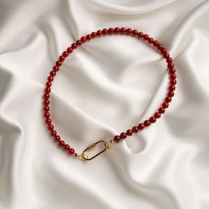 May include: A red beaded necklace with a gold-coloured clasp, displayed on a white satin surface. The necklace features small, round beads. The clasp is a simple, elongated oval shape.