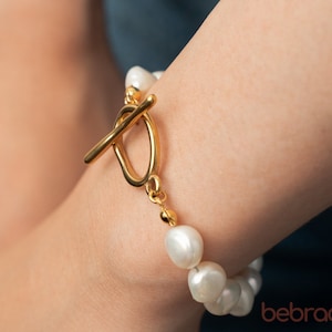 May include: A gold-toned bracelet featuring large, irregular white pearls. The bracelet has a gold toggle clasp. The pearls are strung together, creating a classic and elegant jewelry piece. The bracelet is shown on a person's wrist.