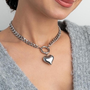 Peut inclure: Un collier en chaîne argentée avec un grand pendentif en forme de cœur. Le collier est porté par une personne avec un pull gris.