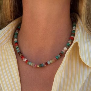 May include: A beaded necklace featuring a repeating pattern of small, round, multi-colored beads in shades of red, turquoise, white, and brown, accented by a single small, polished gold-toned bead centered at the front of the strand.
