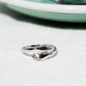 May include: A silver ring featuring two hands clasped together, representing love and unity. The ring is on a white surface.