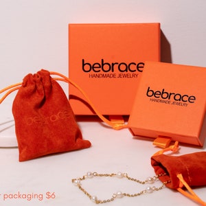 Op de afbeelding: Twee oranje dozen met de tekst "bebrace HANDMADE JEWELRY" en twee rode fluwelen trekkoordzakjes. De dozen en zakjes worden gebruikt voor cadeauverpakkingen.