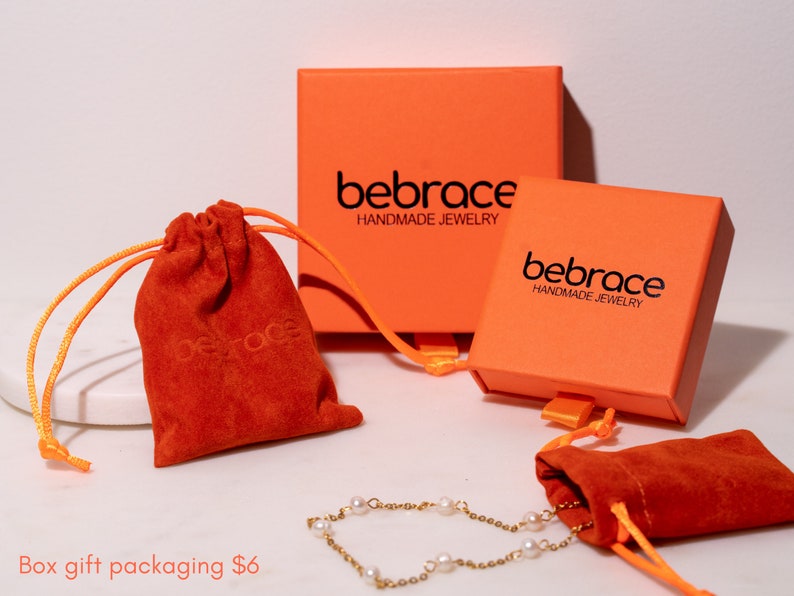 Op de afbeelding: Twee oranje dozen met de tekst "bebrace HANDMADE JEWELRY" en twee rode fluwelen trekkoordzakjes. De dozen en zakjes worden gebruikt voor cadeauverpakkingen.