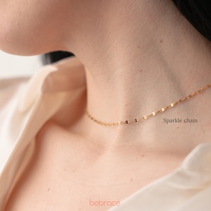 Op de afbeelding: Een gouden ketting met een delicaat glinsterend kettingontwerp. De ketting wordt gedragen door een persoon met een wit shirt. De tekst "Sparkle chain" is zichtbaar in de afbeelding.