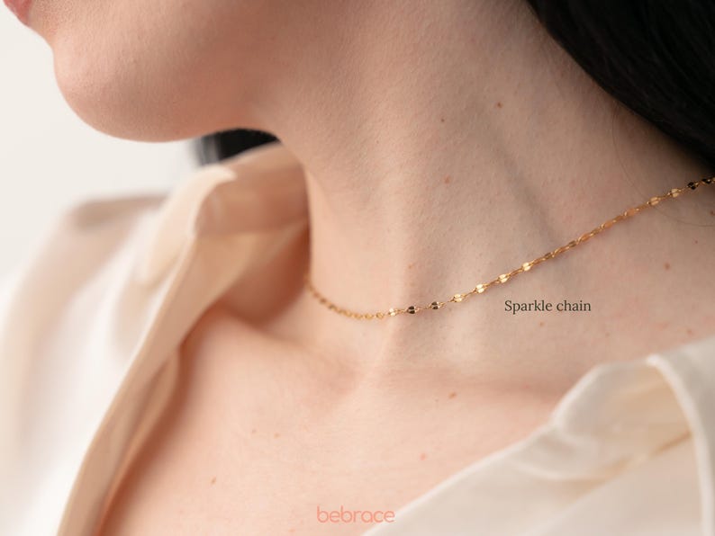 Op de afbeelding: Een gouden ketting met een delicaat glinsterend kettingontwerp. De ketting wordt gedragen door een persoon met een wit shirt. De tekst "Sparkle chain" is zichtbaar in de afbeelding.