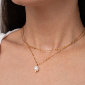 May include: A gold chain necklace with a single pearl pendant. The necklace is layered with two chains.