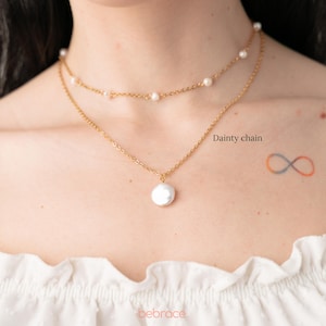 Op de afbeelding: Een gouden ketting met een grote parel hanger en een kleinere parelketting eroverheen gelegd. De tekst "Dainty chain" staat op de afbeelding.