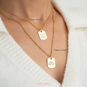 Op de afbeelding: Twee gouden kettingen met bijpassende rechthoekige hangers. De bovenste ketting is een ankerketting met een hanger waarop "Made of Love, Laughter, and Sweet Dreams" staat. De onderste ketting is een figaro-ketting met een hanger waarop "Stay Hungry" staat.