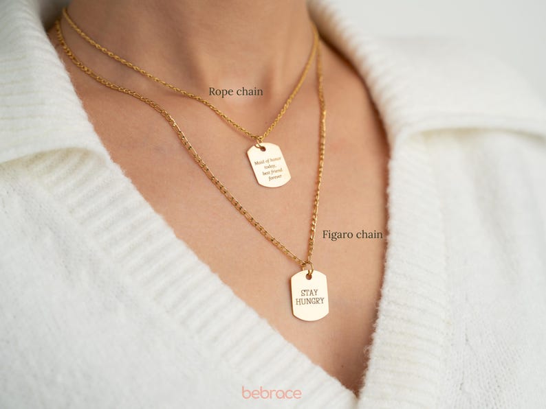 Op de afbeelding: Twee gouden kettingen met bijpassende rechthoekige hangers. De bovenste ketting is een ankerketting met een hanger waarop "Made of Love, Laughter, and Sweet Dreams" staat. De onderste ketting is een figaro-ketting met een hanger waarop "Stay Hungry" staat.