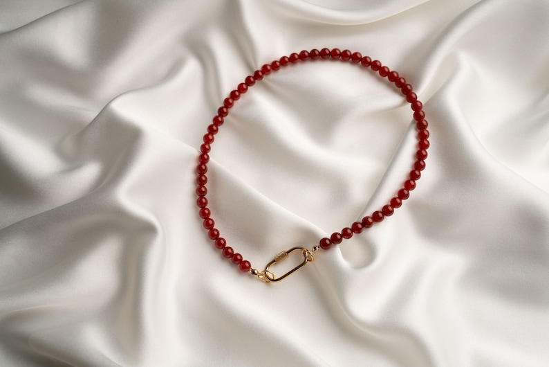 May include: A red carnelian beaded necklace with a gold clasp. The necklace is on a white satin background.