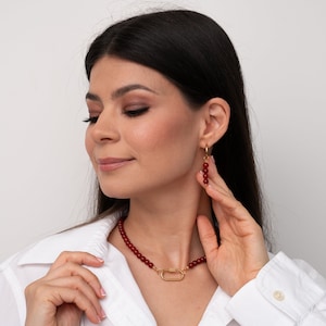 May include: A woman wearing a white shirt with a red beaded necklace and matching earrings. The necklace has a gold clasp in the shape of a rectangle.
