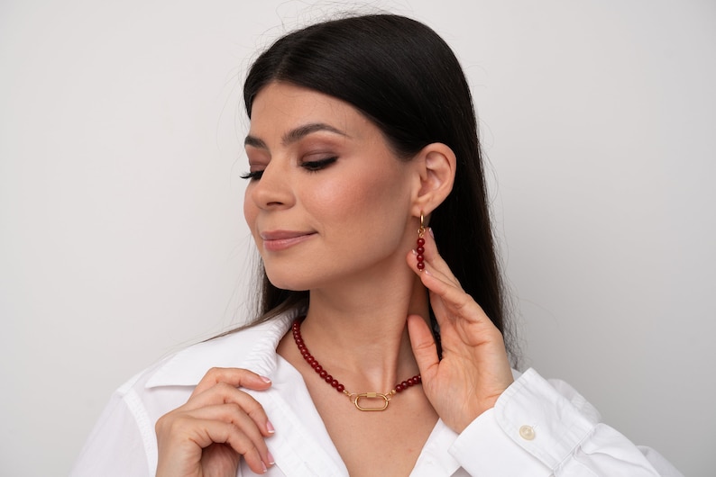 May include: A woman wearing a white shirt with a red beaded necklace and matching earrings. The necklace has a gold clasp in the shape of a rectangle.