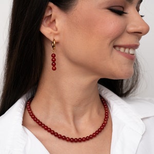 May include: A red carnelian necklace and earrings set. The necklace is made of small, round beads and the earrings are dangle earrings with a gold hoop and red carnelian beads.